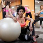 young ethnic woman with fit ball sitting on floor while training in modern gym