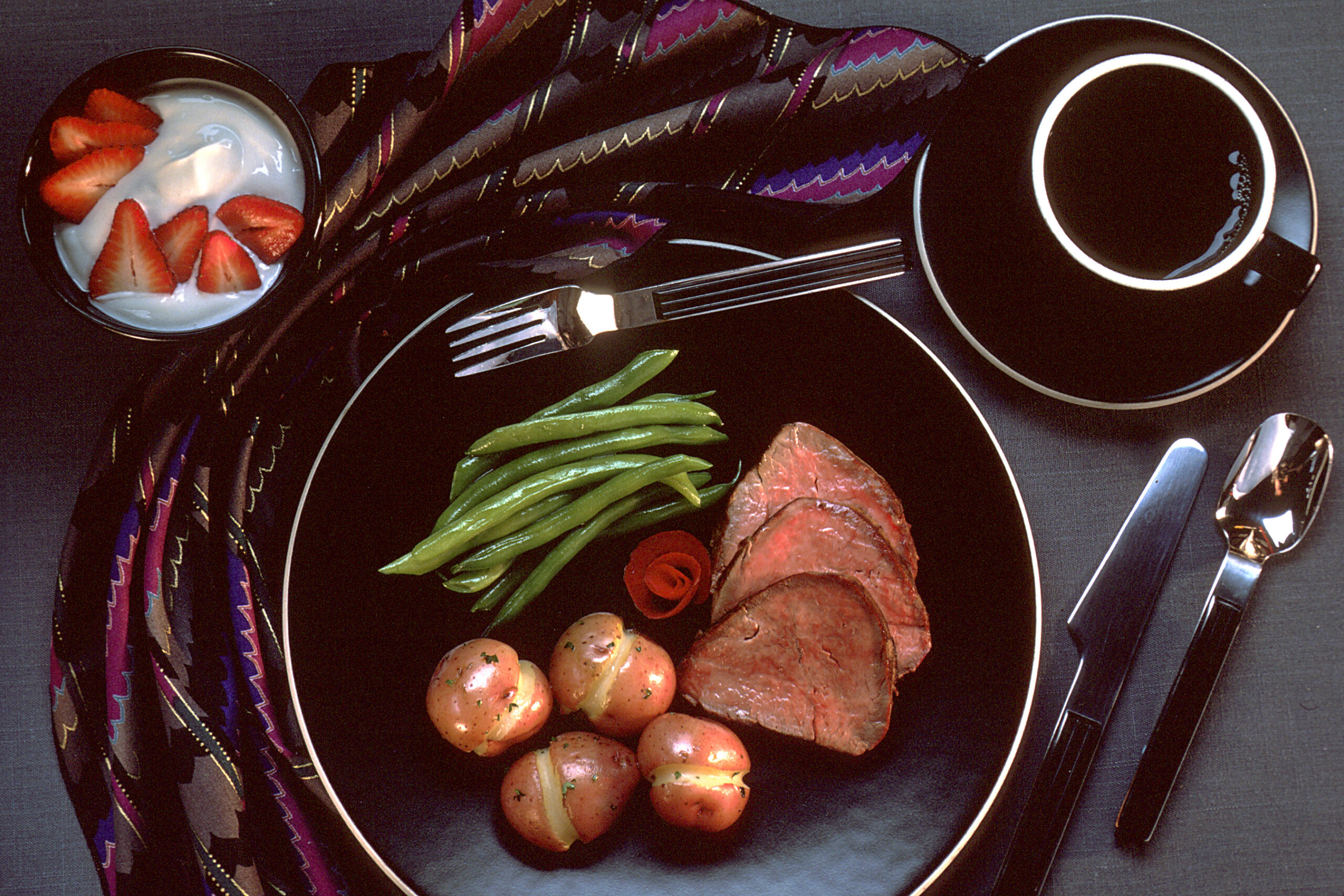 A dinner plate with roast beef, green beans, potatoes, strawberries, and a mug on a dark tabletop